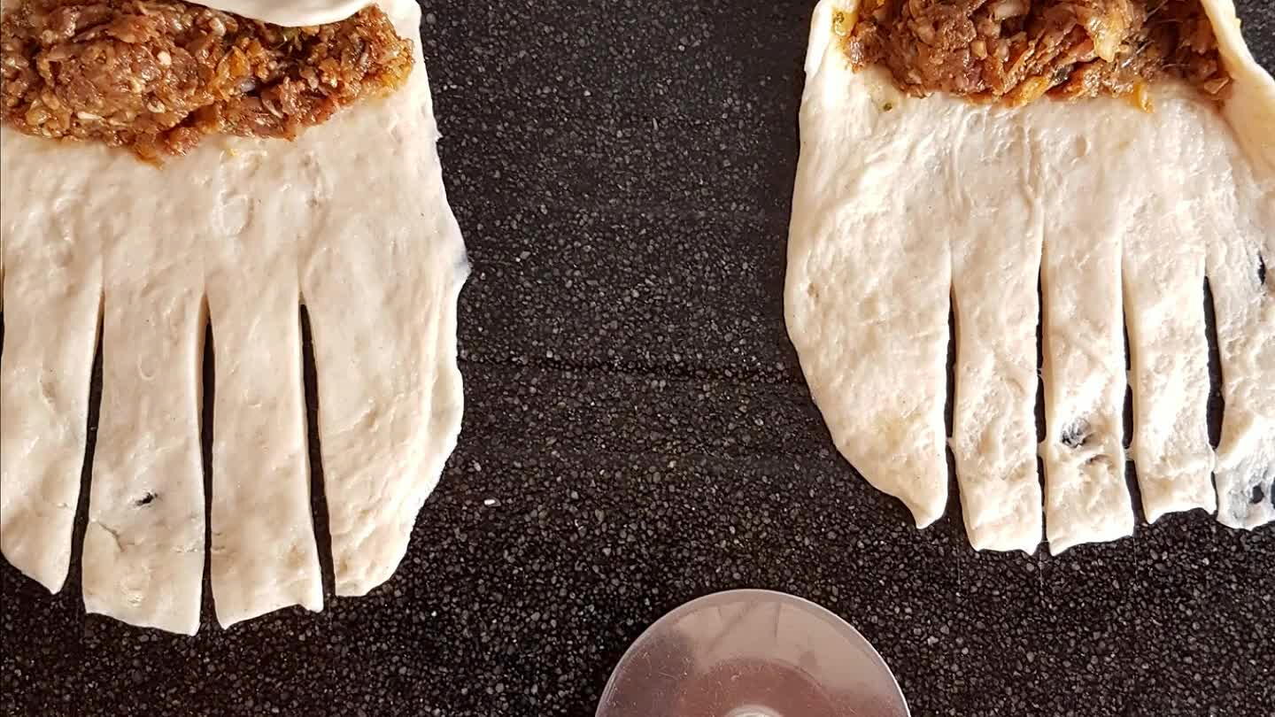 balletje plat naar een wat langwerpige vorm. Schep er een lepeltje gehakt op en snij verticale streepjes aan de onderkant. Rol het broodje op en leg op de bakplaat.
